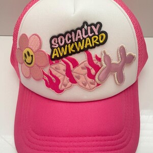May include: A white and pink trucker hat with a black patch that says "Socially Awkward". The hat has a pink flower patch with a smiley face, a pink and white flame patch, and a pink balloon animal patch.