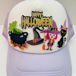 May include: A lavender and white Halloween-themed trucker hat. The front panel features the words "Happy Halloween" with cartoon illustrations of a witch, cauldron, cat, and witch's hat. The hat has a mesh back and a curved brim.
