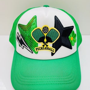 May include: A green and white trucker hat with a black star, a green star with sequins, a green and yellow pickleball paddle, and a green flower patch. The hat says "Pickleball" on the front.