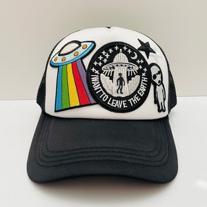 May include: A white trucker hat with black mesh back. The hat features embroidered patches of a UFO, a rainbow, a star, and a patch that says "I want to leave the earth".