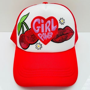 May include: A red and white trucker hat with a red heart patch that says "Girl Power" in pink lettering. The hat also has a red cherry and a red lip patch with sequins.