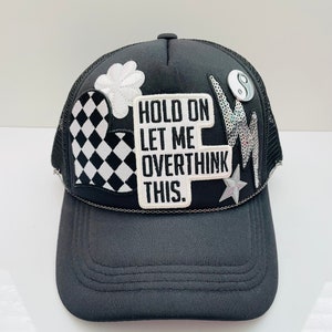 May include: Black trucker hat with a variety of patches, including a checkered heart, a flower, a star, and a patch that says "Hold on let me overthink this."
