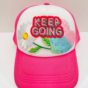 May include: White and pink trucker hat with a pink mesh back. The hat has a white daisy, a pink heart with glitter, and a pink "KEEP GOING" patch.