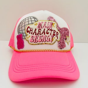 May include: Pink and white trucker hat with a gold chain, a pink sequined patch, a gummy bear patch, a disco ball patch, and a white patch with the text "Main Character Energy".