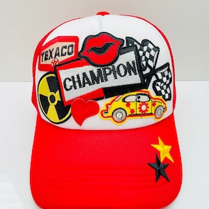 May include: A red and white trucker hat with various patches, including a red lip, a yellow car, a checkered flag, a red heart, a yellow and black radiation symbol, and a black and white "Champion" patch. The hat also has two stars on the back, one gold and one black.