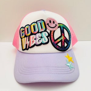 May include: A white and pink trucker hat with a rainbow peace sign patch, a smiley face patch, and the words "GOOD VIBES" in colorful letters. The hat has a purple brim with two star patches.