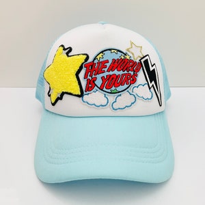 May include: A light blue trucker hat with a white mesh back. The front of the hat features a colorful patch with a star, clouds, a lightning bolt, and the text "THE WORLD IS YOURS".