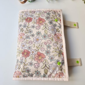 May include: A floral patterned journal cover with a cream background and pink, peach, and blue flowers. The cover has a light pink border, beige straps, and green snap closures. The journal cover is designed to protect and decorate notebooks.