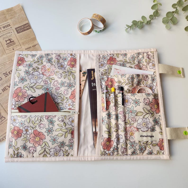May include: Open floral fabric journal cover with multiple pockets and a zipper pocket. The cover is cream-colored with a floral pattern in pink, peach, and green. The journal cover contains a notebook, pens, and washi tape. The label says "Atelierhippie".