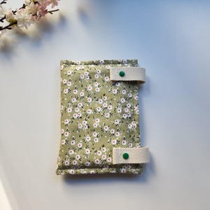 May include: A floral print wallet with a light green background and small white and pink flowers. The wallet has two beige straps with green button closures. The wallet is rectangular and appears to be made of fabric.