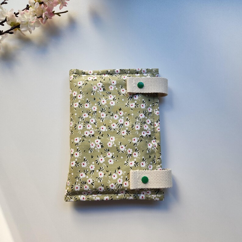 May include: A floral print wallet with a light green background and small white and pink flowers. The wallet has two beige straps with green button closures. The wallet is rectangular and appears to be made of fabric.