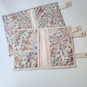 May include: Two floral fabric book covers with a cream background, featuring pink, white, and green flowers. One cover has a zippered pocket and the other has a label that reads "Atelierhippie". Both have cream-colored straps with orange buttons.