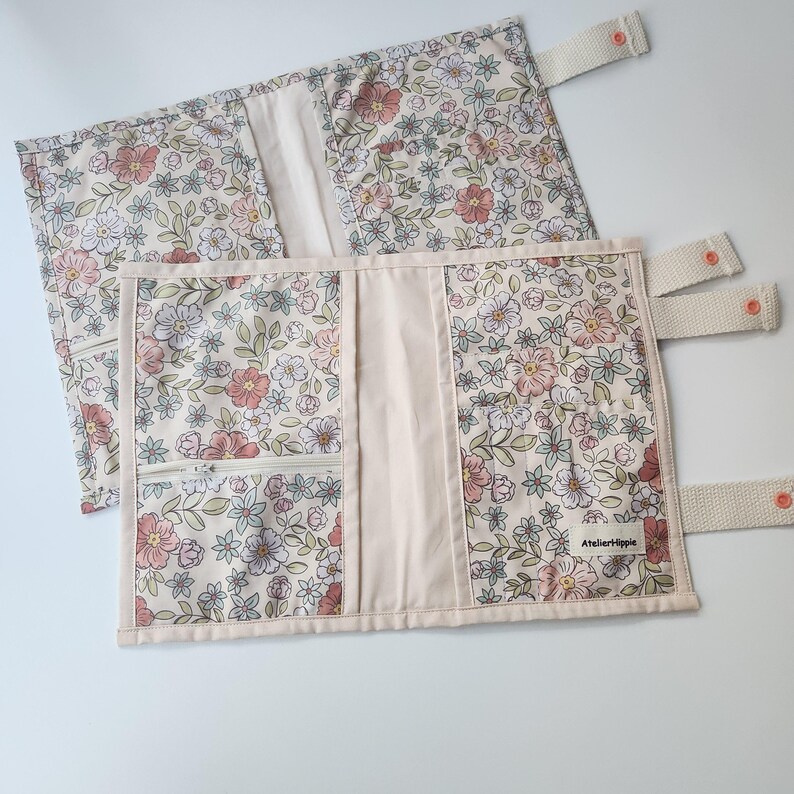 May include: Two floral fabric book covers with a cream background, featuring pink, white, and green flowers. One cover has a zippered pocket and the other has a label that reads "Atelierhippie". Both have cream-colored straps with orange buttons.