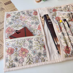 May include: A floral print organizer with multiple pockets and a zipper closure. The fabric features a cream background with pink, peach, blue, and green flowers. A book and pens are visible in the pockets, along with a brown card.