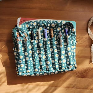 May include: A rectangular fabric pencil case featuring a dark green background with a repeating white daisy pattern. The front of the case has six vertical pockets, each containing a single pen or mechanical pencil of various colors and designs.