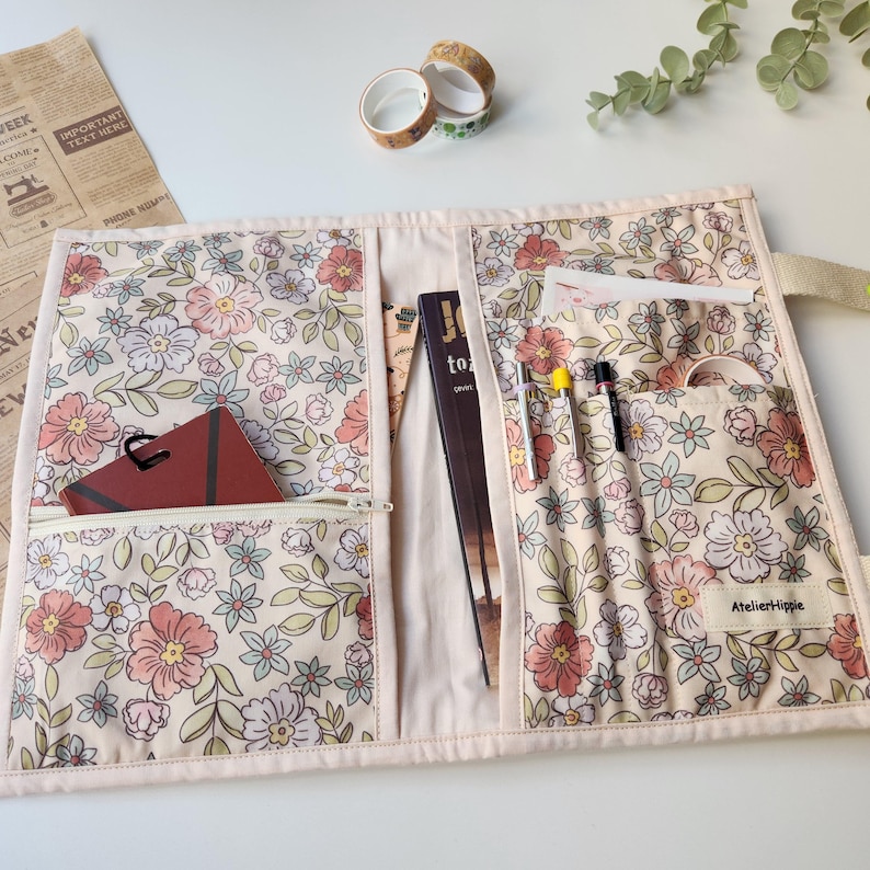 May include: Floral fabric organizer with multiple pockets and a zipper pocket. The organizer is cream-colored with a pink and green floral pattern. It contains a notebook, pens, and a ruler. The brand name "AtelierHippie" is visible on the right side.