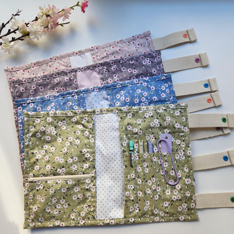 May include: A collection of floral fabric knitting needle cases in shades of green, blue, purple, and pink. Each case has a cream-colored strap with a button closure and interior pockets for tools. A zipper pocket is visible on the front.