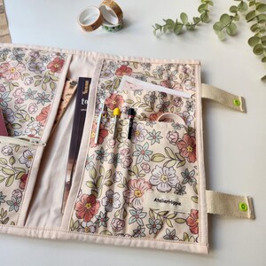 May include: A floral print journal cover with multiple pockets and a zipper closure. The cover is cream-colored with a pattern of pink, peach, and green flowers. It has a beige strap with a green button closure. The label says "AtelierHippie".