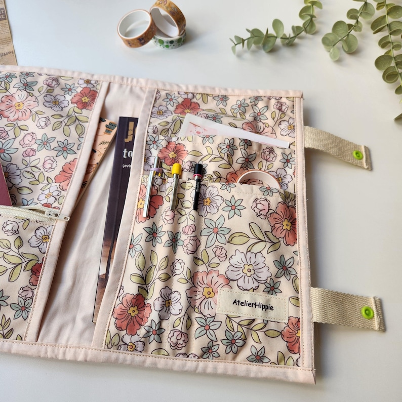May include: A floral print journal cover with multiple pockets and a zipper closure. The cover is cream-colored with a pattern of pink, peach, and green flowers. It has a beige strap with a green button closure. The label says "AtelierHippie".