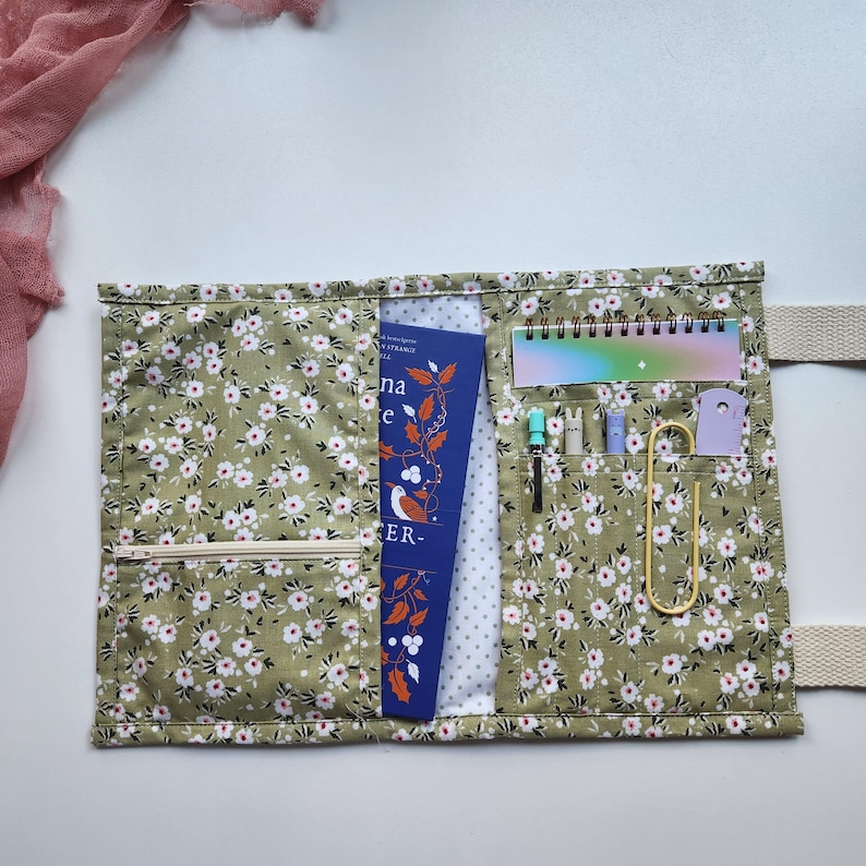 May include: A floral print fabric organizer with a zipper pocket, holding a book, pens, a notepad, and a paperclip. The organizer is olive green with white flowers and has a cream-colored strap.