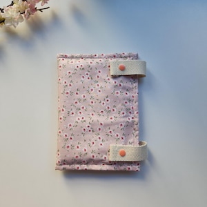 May include: A floral print journal cover in a soft pink hue, adorned with a delicate pattern of tiny white and red flowers. The cover features two beige fabric straps with coral-colored button closures, offering a secure and stylish closure.