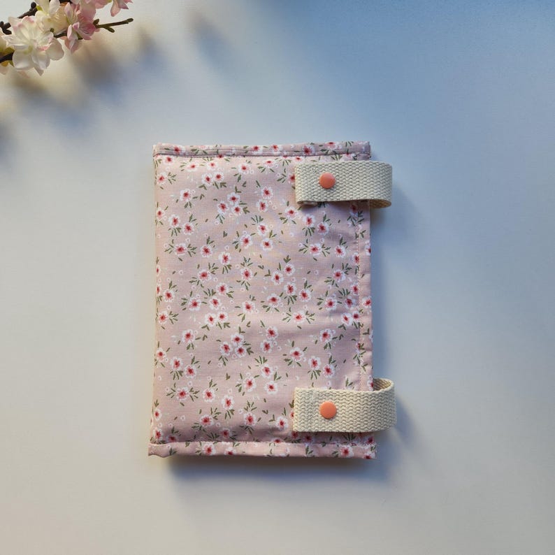 May include: A floral print journal cover in a soft pink hue, adorned with a delicate pattern of tiny white and red flowers. The cover features two beige fabric straps with coral-colored button closures, offering a secure and stylish closure.