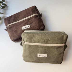May include: Two rectangular zippered pouches, one in dark brown and one in olive green. Each pouch features a light-colored zipper, a textured fabric finish, and a white rectangular fabric label on the front with the text AtelierHippie.