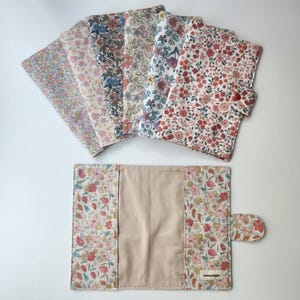 May include: A collection of floral fabric items, including several folded squares and a book cover. The fabrics feature a variety of colorful floral patterns on a white background. The book cover has a beige interior and a button closure.