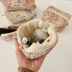 Könnte beinhalten: Eine kleine Stofftasche mit Blumendruck und Reißverschluss. Die Tasche ist offen und mit verschiedenen Toilettenartikeln gefüllt, darunter eine Glasflasche Parfüm, eine kleine Tube Lotion und eine Tropfflasche.