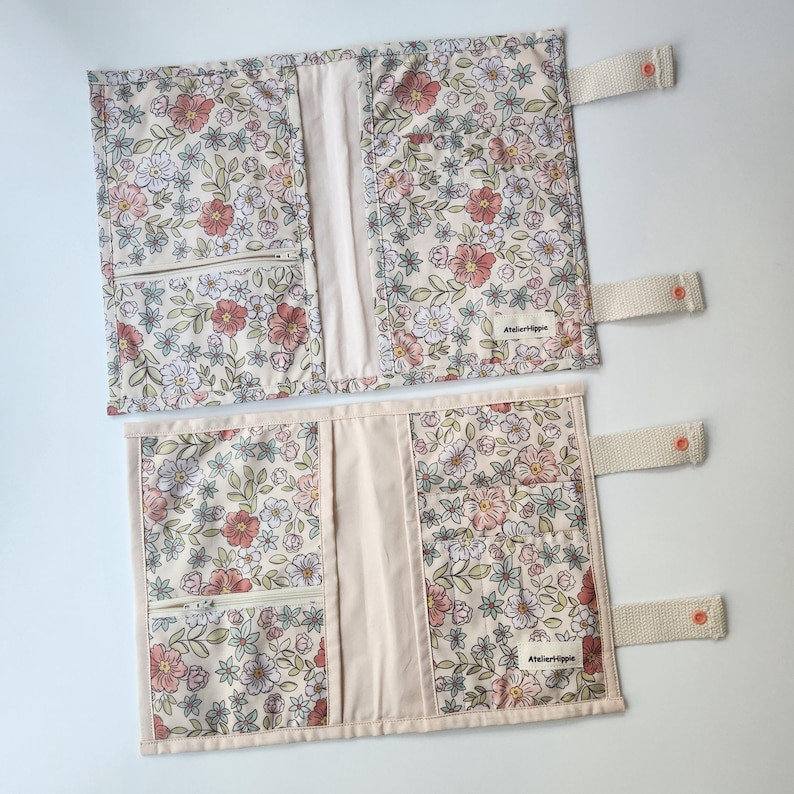 May include: Two floral fabric book covers with a cream background, featuring pink and white flowers and green leaves. Each cover has a cream-colored interior, a zippered pocket, and a beige strap with a coral button. The text "Atelierhippie" is visible.