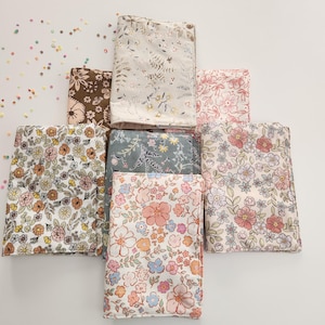 May include: A collection of fabric items, possibly book covers or small pouches, arranged in a starburst pattern. The fabrics feature floral patterns in various colors, including pink, peach, and brown, with a white background. Some have a light beige background with floral designs.