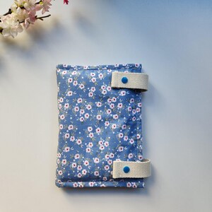 May include: A blue floral fabric case with white straps and blue button closures. The case is covered in a pattern of small white and pink flowers. The straps are a light beige color.