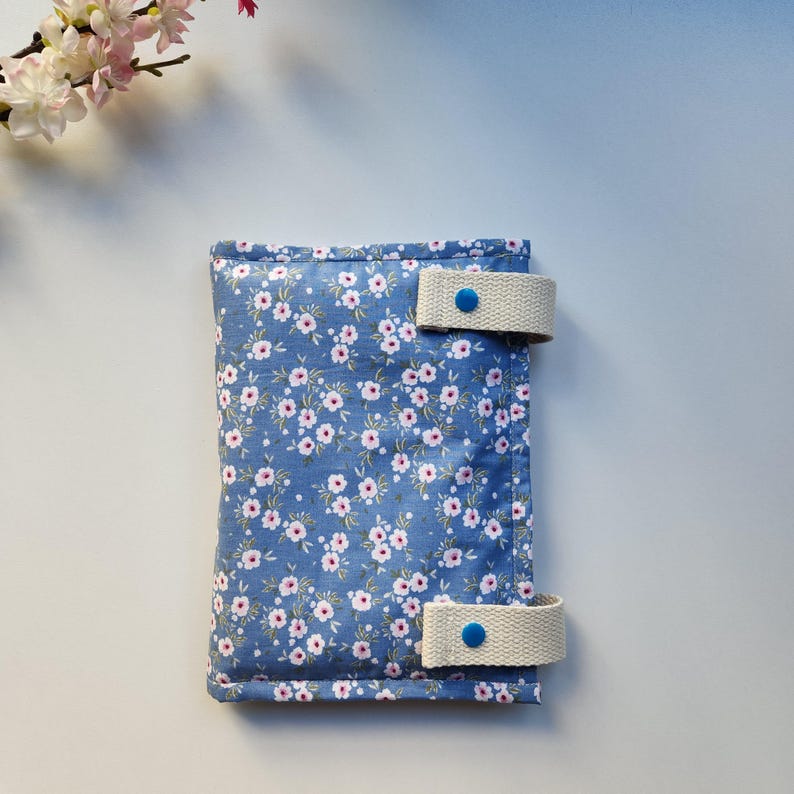 May include: A blue floral fabric case with white straps and blue button closures. The case is covered in a pattern of small white and pink flowers. The straps are a light beige color.