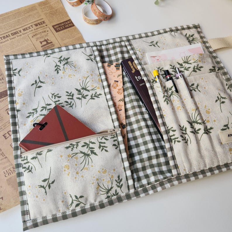 May include: Open fabric organizer with a floral and gingham pattern. The organizer has multiple pockets holding a notebook, pens, and a small book. A zippered pocket holds a red notebook. The text "to" is visible on the spine of a book.