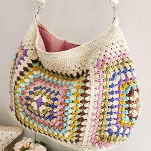 May include: A handmade crochet shoulder bag in a granny square pattern. The bag features a cream-colored top and strap, with a colorful patchwork design in shades of brown, purple, pink, blue, and yellow. The interior lining is a soft pink fabric.