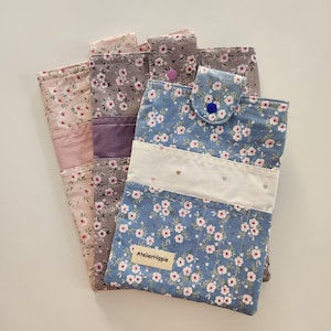 May include: Three rectangular fabric pouches featuring floral patterns in blue, grey, and pink. Each pouch has a horizontal band, a snap-button closure, and a small beige label with the text AtelierHippie stitched onto the front surface.