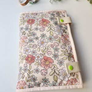 May include: A floral print journal cover with a cream background and pink, peach, and blue flowers. The cover has a beige strap with a green button closure. Decorative washi tape is visible in the background.