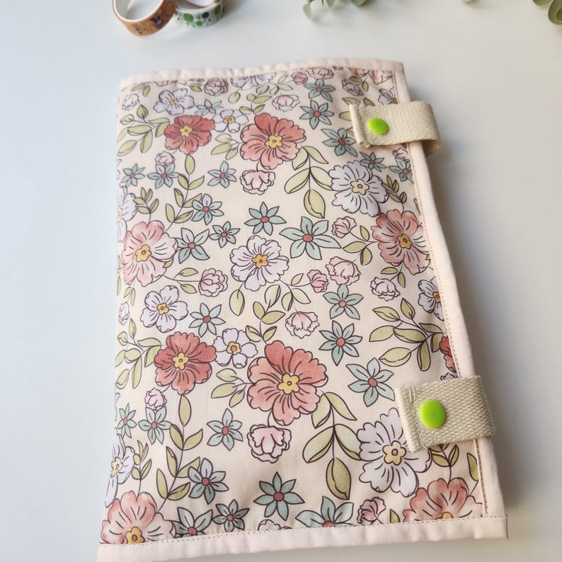 May include: A floral print journal cover with a cream background and pink, peach, and blue flowers. The cover has a beige strap with a green button closure. Decorative washi tape is visible in the background.