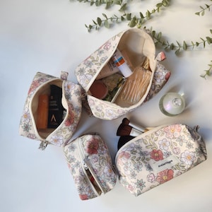 Peut inclure: Quatre trousses zippées en tissu crème avec un motif floral dans les tons rose, bleu et vert. Les trousses sont de tailles variées, présentées ouvertes et fermées, contenant divers articles cosmétiques et une étiquette AtelierHippie.