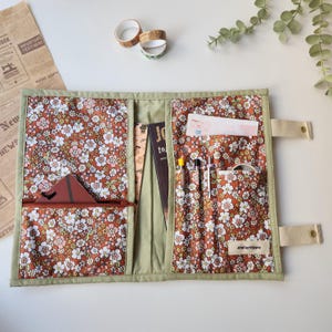 May include: Open floral print organizer with multiple pockets and a zipper pouch. The interior features a brown and white floral pattern, with a green border. The organizer holds pens, a notebook, and other stationery items. The label says "Atelierhippie".