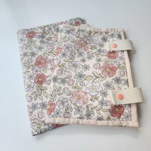 May include: Two floral patterned fabric items with a cream background, featuring pink, white, and blue flowers with green leaves. Each item has a cream-colored border and a cream strap with a pink button closure. The items are likely fabric organizers or covers.