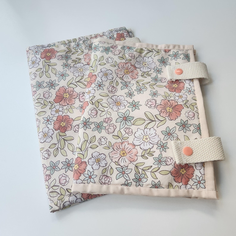 May include: Two floral patterned fabric items with a cream background, featuring pink, white, and blue flowers with green leaves. Each item has a cream-colored border and a cream strap with a pink button closure. The items are likely fabric organizers or covers.