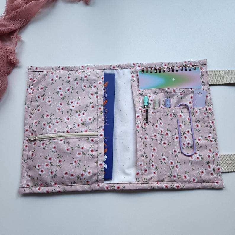 May include: Open floral print notebook cover in pink with white and red flowers. The cover has a zipper pocket, a blue notebook, and pockets holding pens, a paperclip, and a small notepad. A cream-colored strap is attached.