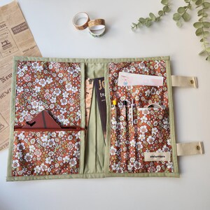 May include: Open, floral-patterned organizer with multiple pockets and compartments. The interior features a brown fabric with white and pink flowers. A zippered pocket, pen holders, and a small pocket are visible. The organizer is trimmed with green fabric and has beige straps.