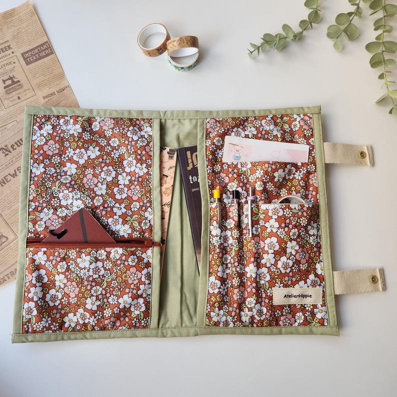 May include: Open, floral-patterned organizer with multiple pockets and compartments. The interior features a brown fabric with white and pink flowers. A zippered pocket, pen holders, and a small pocket are visible. The organizer is trimmed with green fabric and has beige straps.