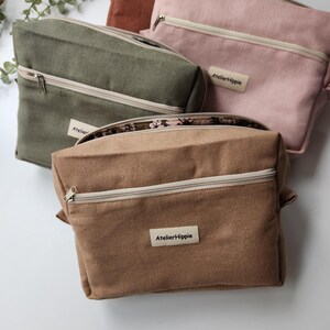 May include: Three rectangular zippered pouches in olive green, dusty pink, and tan. Each pouch features a front exterior pocket with a beige zipper, a cream-colored fabric label with the text AtelierHippie, and a floral patterned interior lining.