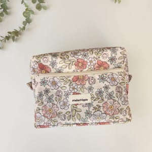May include: A floral print cosmetic bag with a cream-colored background. The bag features a zipper closure and is adorned with pink, white, and blue flowers and green leaves. The label reads "AtelierHippie".