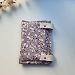 May include: A floral print fabric journal cover in shades of gray and pink. The cover features a repeating pattern of small flowers and is secured with two cream-colored straps and purple button closures. The journal cover is a handmade item.