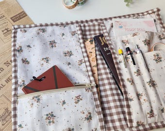 Könnte beinhalten: Ein Stoff-Organizer mit mehreren Taschen, mit einem Blumenmuster auf weißem Hintergrund und braunen Vichy-Akzenten. Er enthält eine Reißverschlusstasche, Stifthalter und ein Notizbuch. Dekorative Klebebandrollen sind sichtbar.