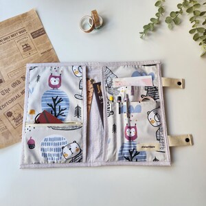 May include: A light gray fabric organizer with a whimsical bear and owl pattern. The organizer has multiple pockets, including a zippered one, and elastic pen holders. Beige straps with snap closures secure the organizer. The text "AtelierHippie" is visible.